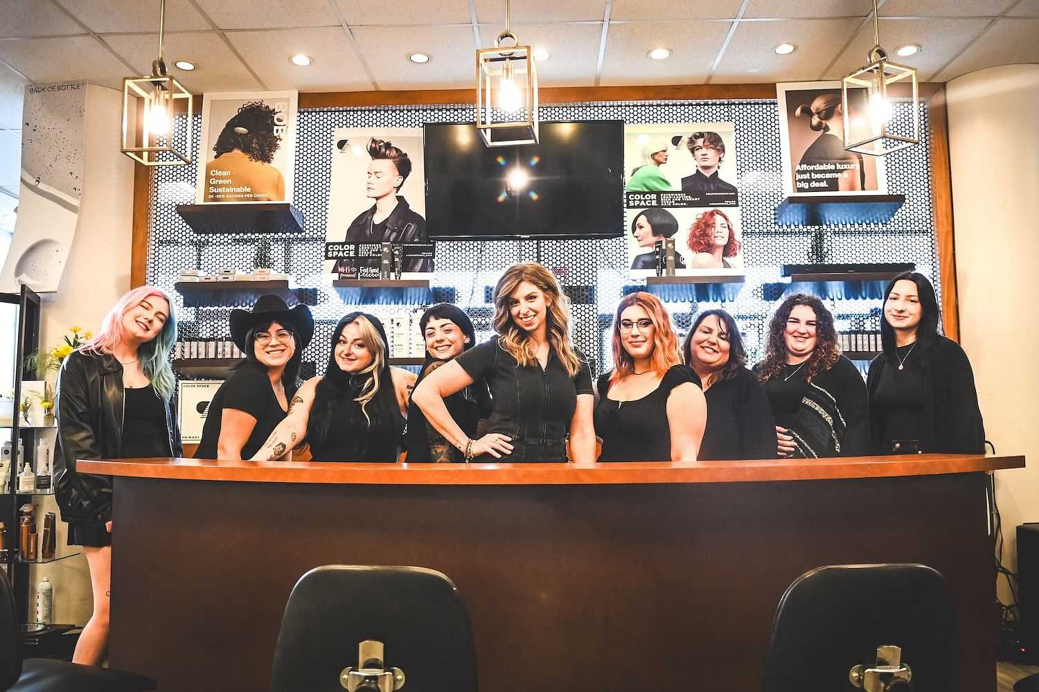 Salon staff smiling behind the reception desk with hairstyle posters in the background.